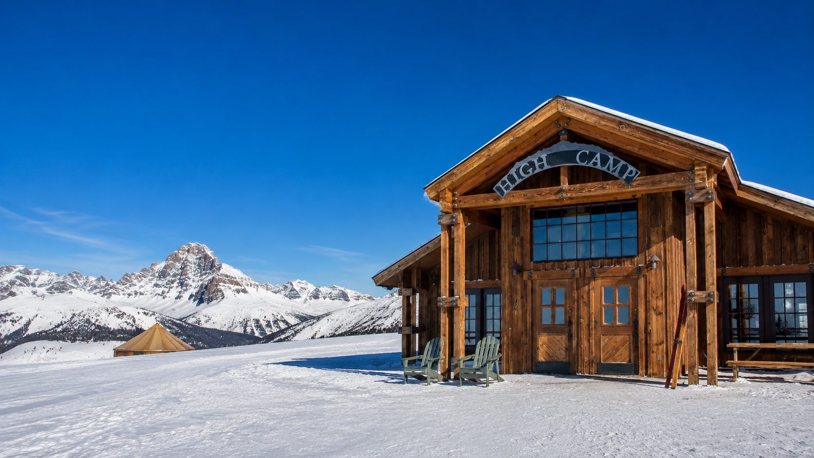 High Camp Warming Hut, a timber-framed lodge at the top of Lift 12 (Prospect Express) at Telluride Ski Resort, with green chairs on the snow out front and a snow-covered peak behind it.